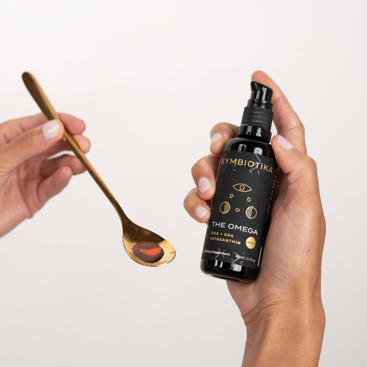 Hand holding a bottle of Cymbiotika The Omega with a spoon containing a droplet of oil on a light gray background