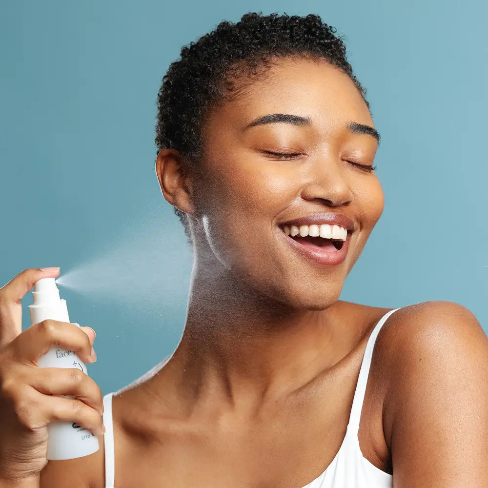 A woman with short curly dark hair smiles while spraying Face Reality Omni Clear Mist onto her face, wearing a white tank top against a solid blue background.