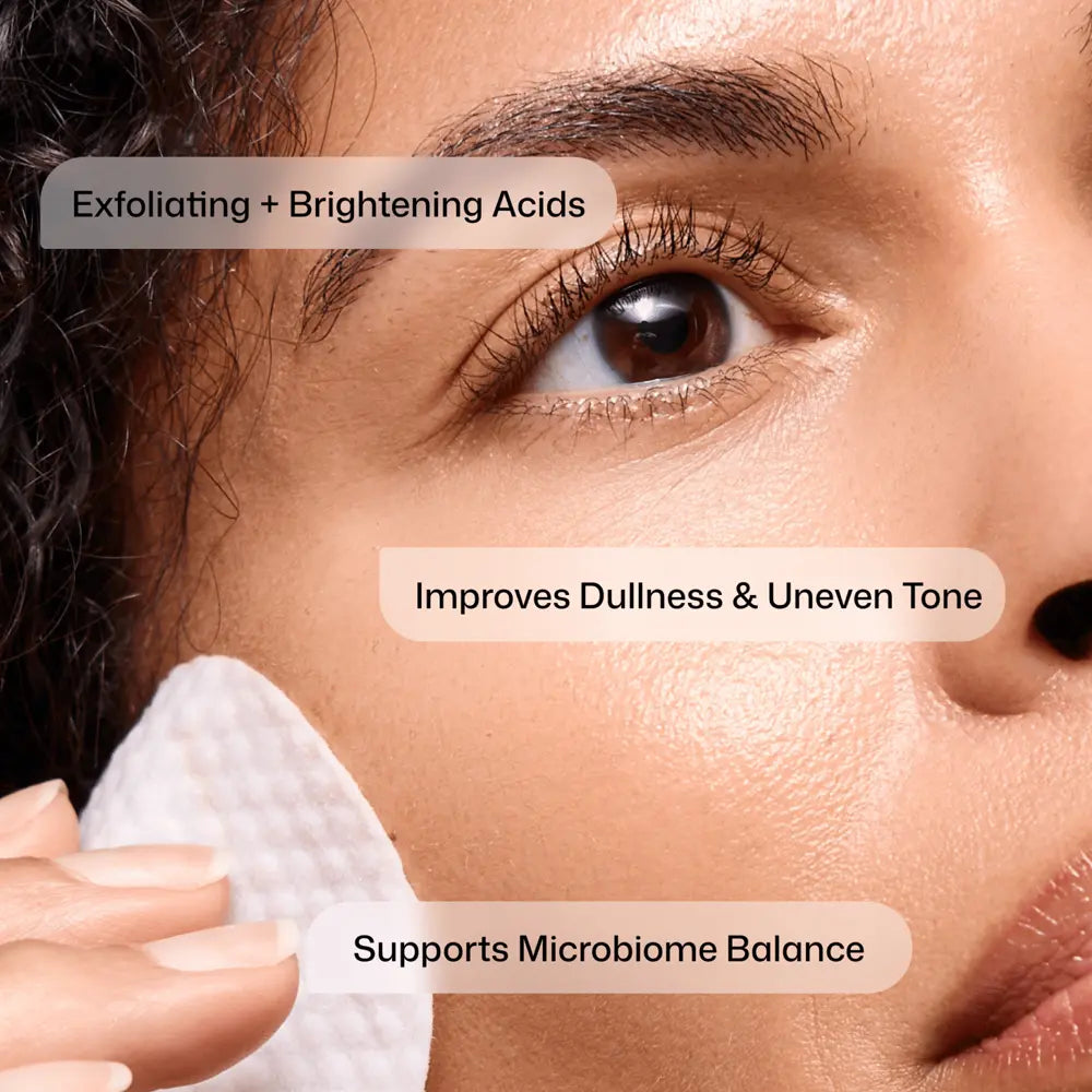 Close-up of a person's face with a Revision Skincare Tone+ Correcting Pad touching the cheek, overlaid with text reading 'Exfoliating + Brightening Acids,' 'Improves Dullness & Uneven Tone,' and 'Supports Microbiome Balance.'.