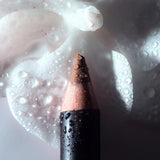 A close-up of Anfisa Skin Ân-Define Ceramide Liner in Burnt Toffee with a warm brown shade, covered in water droplets, set against a white background with a matching white makeup smear.