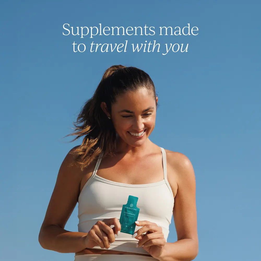 A smiling woman in a white tank top holds a small teal travel-sized supplement pouch against a clear blue sky. White text at the top reads, 'Supplements made to travel with you.