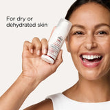A smiling woman holds up a white tube of ella MD Hydration Defense SPF 30 moisturizer against a light beige background, with the text 'For dry or dehydrated skin' on the left.
