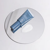 A Face Reality Age Adaptive Clearing Serum, placed diagonally on a round clear glass plate next to a dollop of product texture