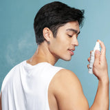 A man in a white tank top sprays Face Reality Omni Clear Mist on his face and body against a solid blue background.