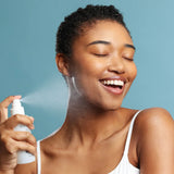 A woman with short curly dark hair smiles while spraying Face Reality Omni Clear Mist onto her face, wearing a white tank top against a solid blue background.