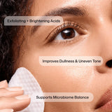 Close-up of a person's face with a Revision Skincare Tone+ Correcting Pad touching the cheek, overlaid with text reading 'Exfoliating + Brightening Acids,' 'Improves Dullness & Uneven Tone,' and 'Supports Microbiome Balance.'.