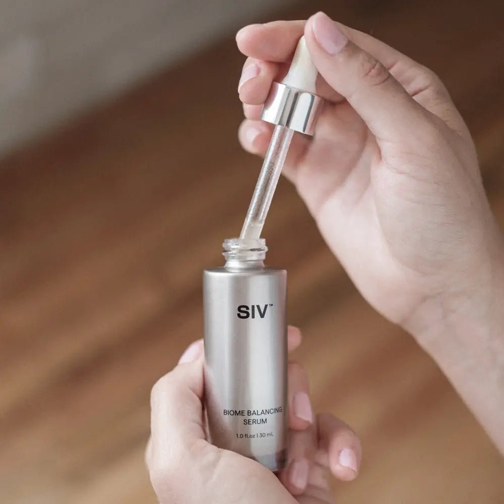 Hand holding a silver SIV serum bottle with dropper against a blurred wooden background