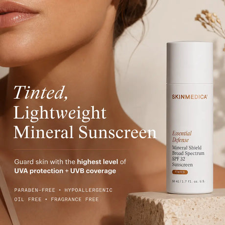 A close-up of a woman's lower face and neck next to a white bottle of  SkinMedica Essential Defense Mineral Shield SPF 32 Tinted. The background is a warm beige with elegant dried floral accents, and the text highlights the product's tinted, lightweight formula.