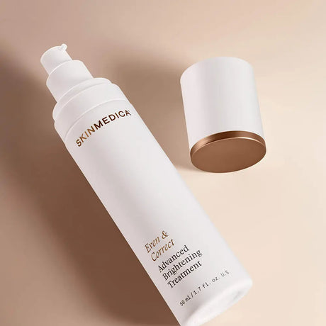 A white pump bottle of SKINMEDICA Even & Correct Advanced Brightening Treatment with gold lettering, shown next to its matching white cap with a metallic gold base on a warm beige background.