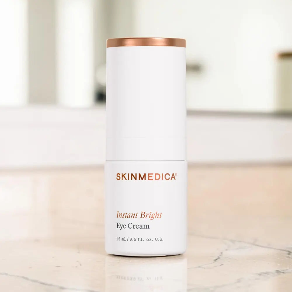 A white cylindrical bottle of SKINMEDICA Instant Bright Eye Cream with a metallic rose gold cap, set on a light marble countertop with a blurred bathroom background.