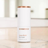 A white cylindrical bottle of SKINMEDICA Instant Bright Eye Cream with a metallic rose gold cap, set on a light marble countertop with a blurred bathroom background.