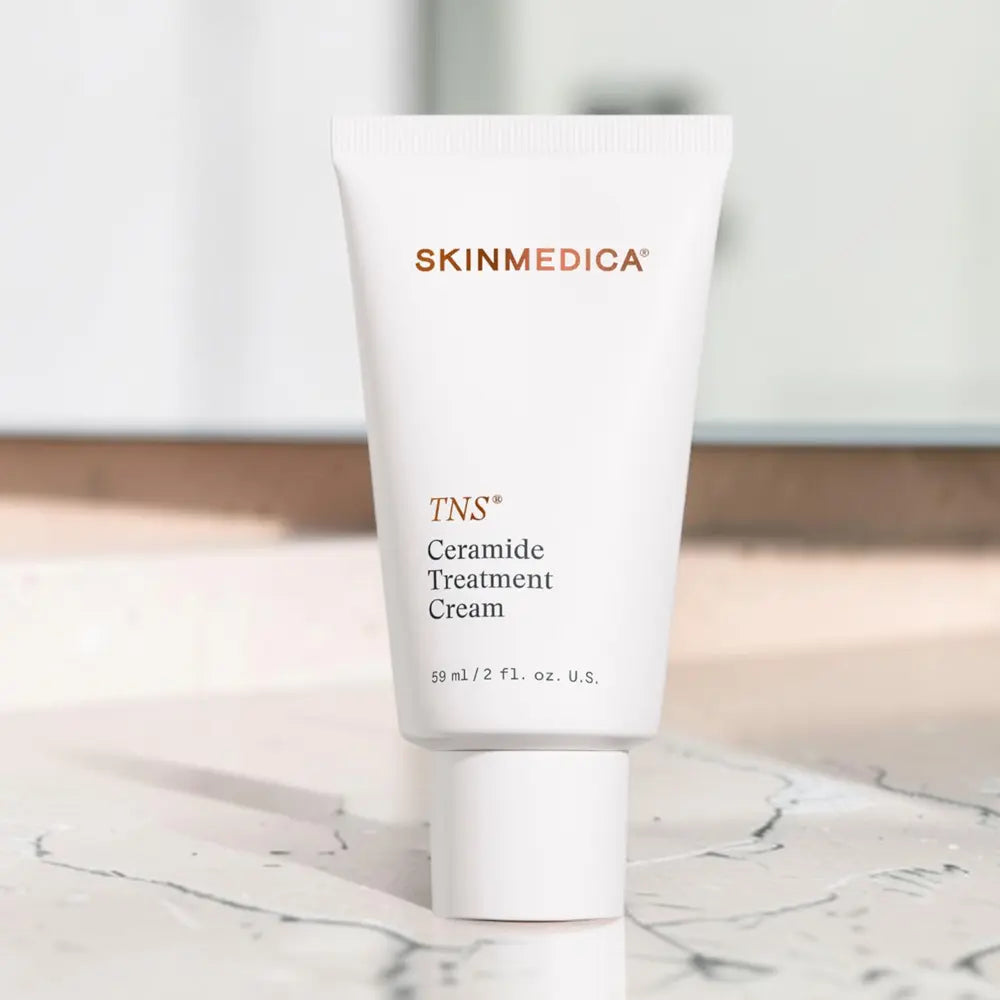 A white squeeze tube of SSkinMedica TNS Ceramide Treatment Cream standing upright on a light marble surface with a softly blurred indoor background.