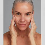 A close-up portrait of a woman with gray hair, applying Adaptogenic Cleanser to her face with both hands against a solid gray background.