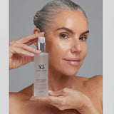 A woman with silver hair pulled back holds a bottle of Young Goose Adaptogenic Cleanser against a neutral gray background.