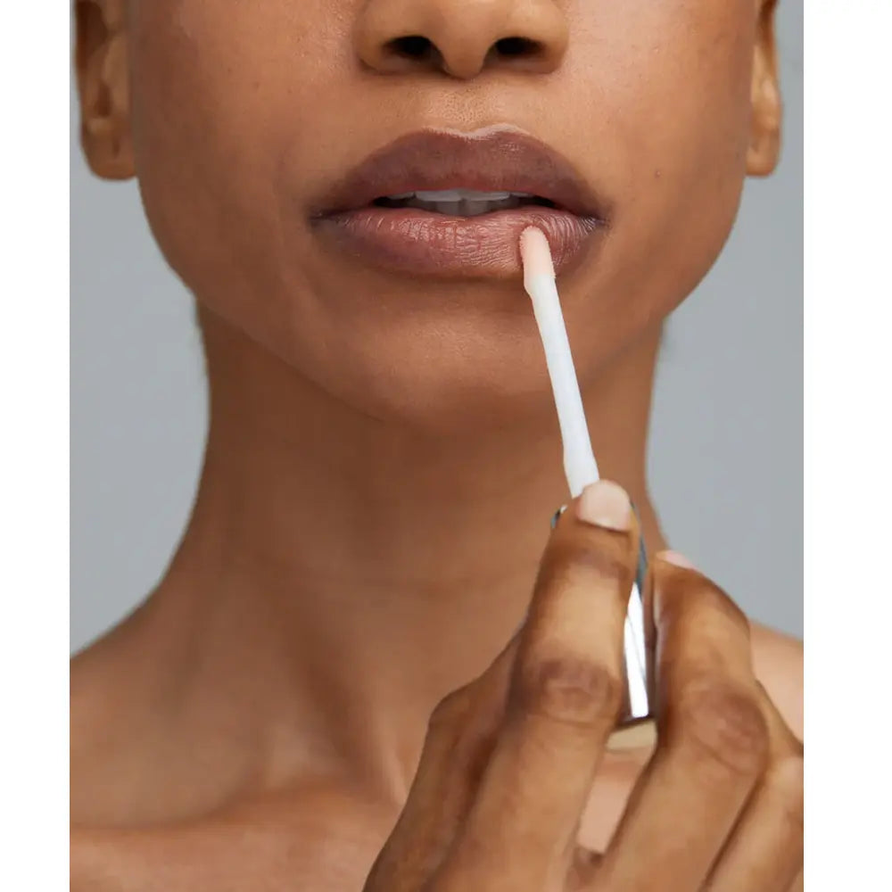 Close-up of a person applying a Young Goose Adaptogloss Anti-Aging Plumper with a doe-foot applicator, shown against a plain gray background.