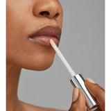 Close-up of a person applying Young Goose Adaptogloss Anti-Aging Plumper with a white doe-foot applicator, against a plain gray background.