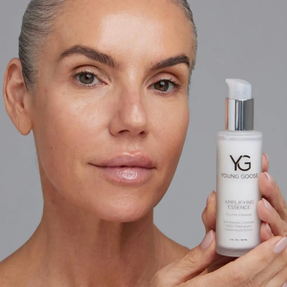 A close-up of a woman with clear, radiant skin holding a white pump bottle of Young Goose Amplifying Essence against a neutral gray background.
