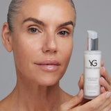 A close-up of a woman with clear, radiant skin holding a white pump bottle of Young Goose Amplifying Essence against a neutral gray background.