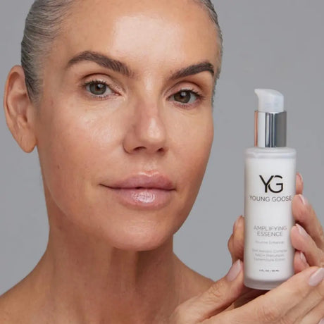 A close-up of a woman with clear, radiant skin holding a white pump bottle of Young Goose Amplifying Essence against a neutral gray background.