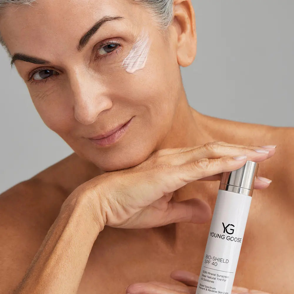 A close-up of a woman applying or demonstrating a skincare product, with a white tube labeled Young Goose Bio-Shield SPF 40 in her hand against a neutral gray background.