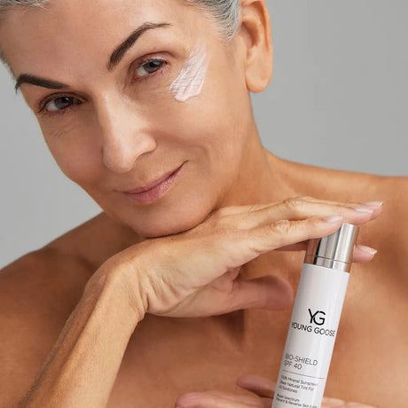A close-up of a woman applying or demonstrating a skincare product, with a white tube labeled Young Goose Bio-Shield SPF 40 in her hand against a neutral gray background.