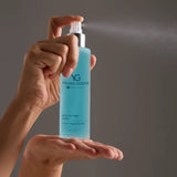 A person's hands holding and spraying a bottle of Young Goose Blue Peptide Spray against a solid gray background.