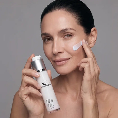 A woman with dark hair pulled back applies a white cream to her cheek while holding a white and silver Young Goose Care pump bottle against a solid gray studio background.