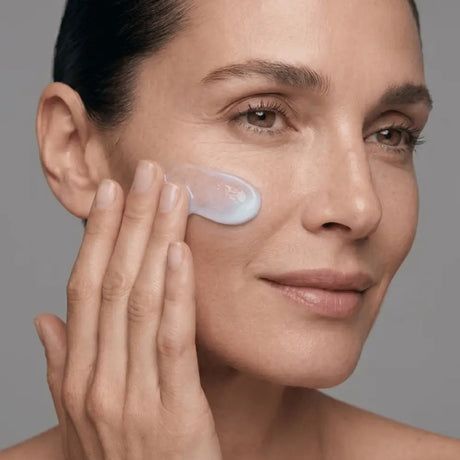Close-up of a woman applying Young Goose CARE to her cheek with her finger against a solid gray background.