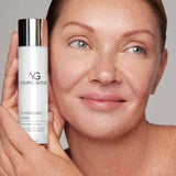 A close-up of a woman holding a white Young Goose Hyperbaric Mask bottle next to her face against a neutral gray background.
