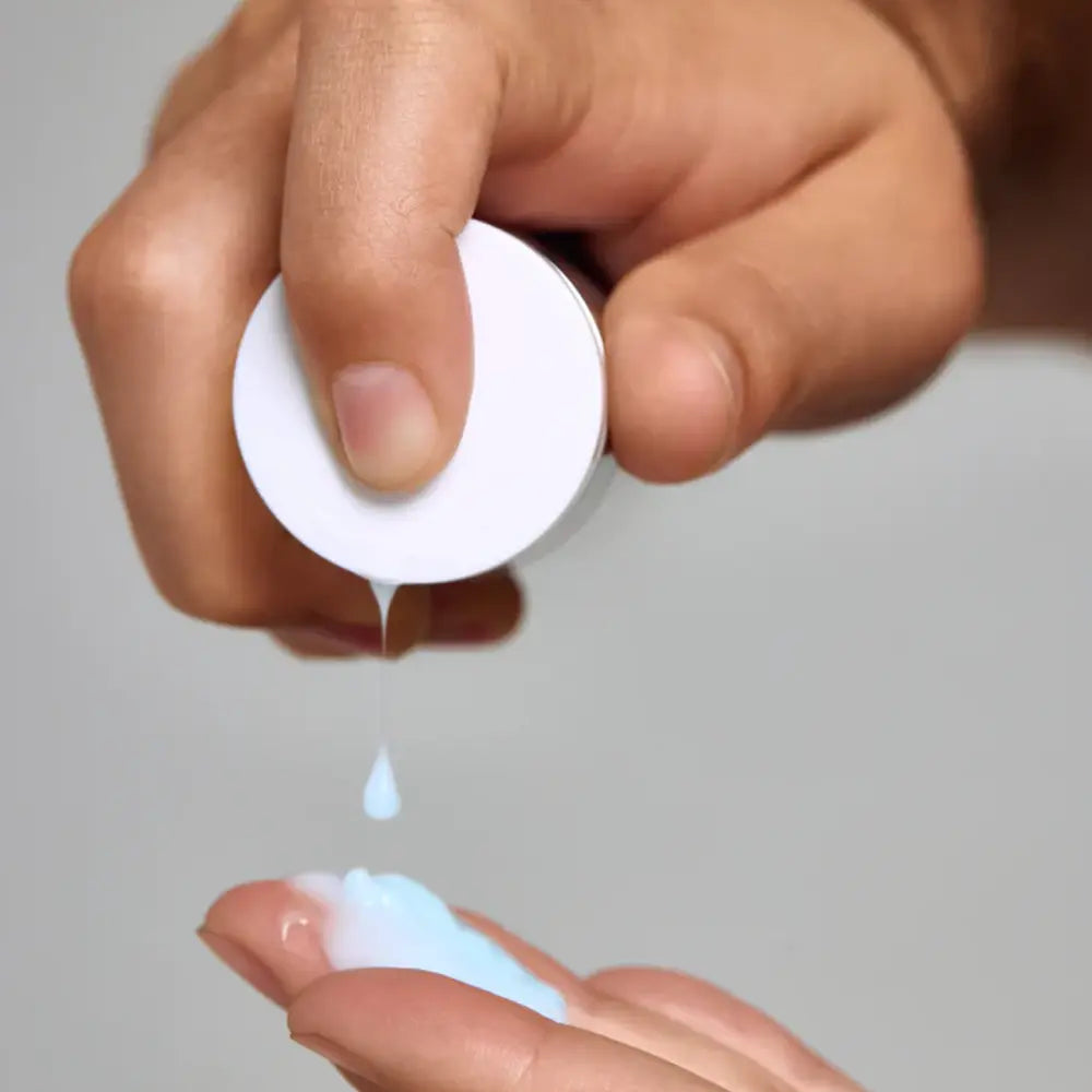 A close-up of a hand squeezing Young Goose L.A.D.R. Serum bottle to dispense the productproduct, with a second hand receiving a small droplet above a larger pool of the product on a neutral light gray background.