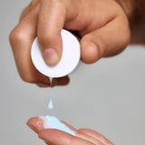 A close-up of a hand squeezing Young Goose L.A.D.R. Serum bottle to dispense the productproduct, with a second hand receiving a small droplet above a larger pool of the product on a neutral light gray background.