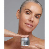 A woman with slicked-back gray hair holds a Young Goose NiaPolish jar near her chest while applying cream with her finger. The background is a solid neutral gray.