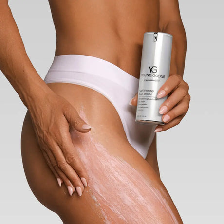 A close-up of a model's hip and thigh applying a pinkish cream while holding a silver can of Young Goose Youth Firming Body Cream against a light gray background.